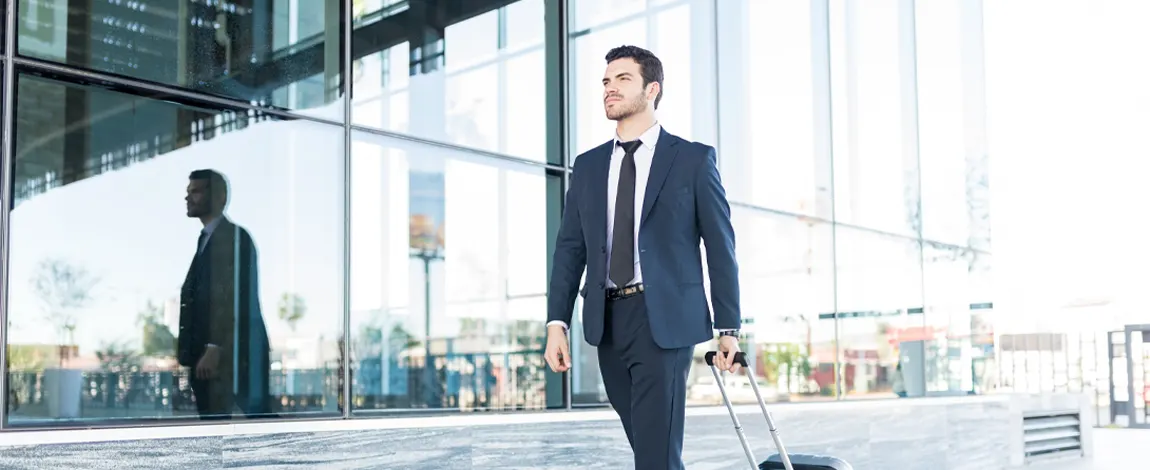 Hombre de negocios caminando con una maleta de viaje frente a un edificio de cristal moderno