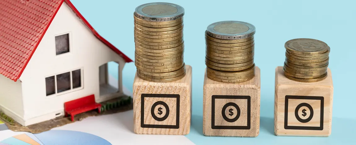 Maqueta de una casa junto a tres pilas de monedas, de mayor a menor, sobre cubos de madera